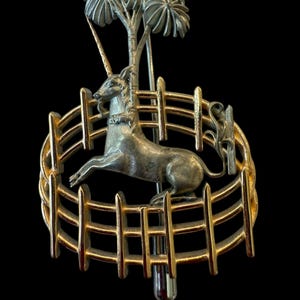 May include: A vintage silver and gold-tone brooch featuring a unicorn leaping within a fence, with a tree in the background. The brooch has a detailed design and a pin closure on the back. The overall theme is fantasy and mythical creatures.