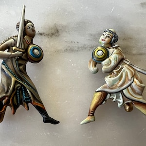 May include: Two ceramic figurines of people in medieval garb, one holding a sword and shield, the other holding a sword and a round shield. The figurines are painted in shades of brown, gold, and blue.