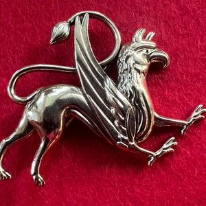 May include: A silver-toned griffin brooch with detailed wings and a fierce expression. The griffin is a mythical creature with the head and wings of an eagle and the body of a lion.
