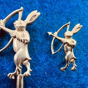 以下が含まれることがあります： 弓と矢を持った2つの銀色のウサギの置物。ウサギは直立しており、詳細な特徴があります。背景は質感のある青い表面です。1つの置物はもう1つよりも大きいです。
