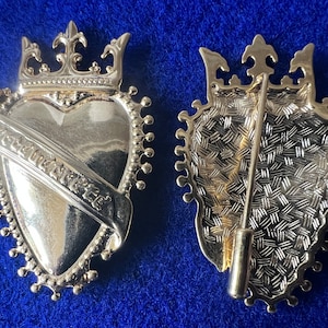 May include: A silver-toned brooch in the shape of a heart with a crown on top. The heart has a banner that reads "Forever" and is surrounded by a beaded border. The back of the brooch shows a pin and a textured design.