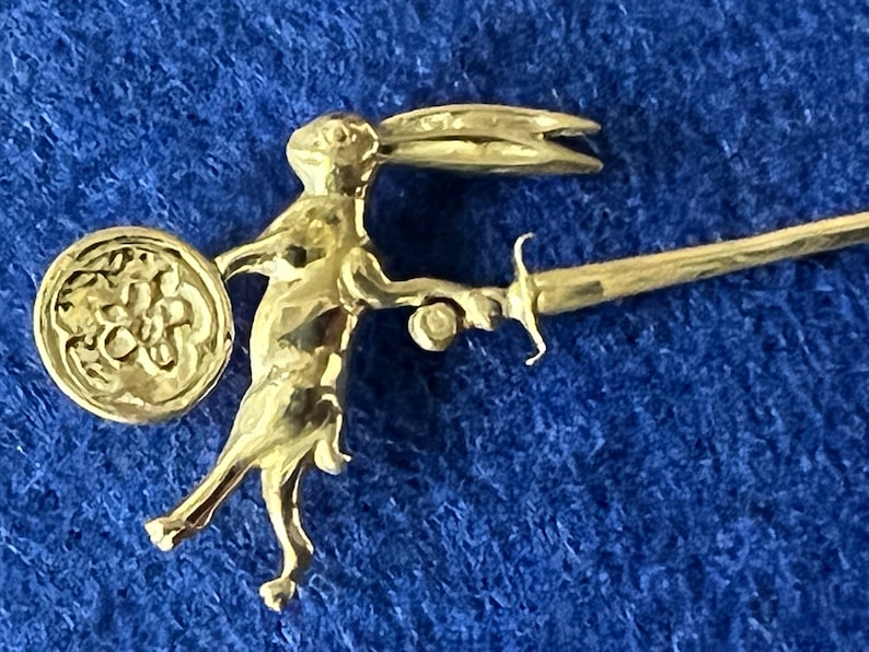 May include: A gold-colored rabbit figurine holding a sword and a shield. The rabbit is standing on its hind legs and has its ears up. The shield is round and has a floral design.