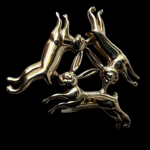 May include: A gold-coloured metal brooch depicting four leaping hares in a circular arrangement. The hares are shown mid-leap, with their ears and legs extended, creating a dynamic and elegant piece of jewellery.