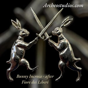 以下が含まれることがあります： 銀色のウサギの置物2つ。それぞれが剣を持ち、戦闘態勢をとっている。画像には「Archeostudios.com」と「Bunny Incrosa - after Fiore dei Liberi」の文字が含まれている。背景は暗く、ぼやけている。