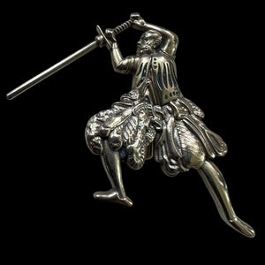 Puede incluir: Una figurita de plata de una persona con una falda fluida que sostiene una espada levantada por encima de su cabeza.