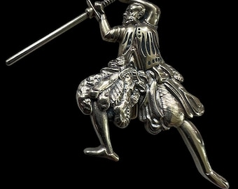 Puede incluir: Una figurita de plata de una persona con una falda fluida que sostiene una espada levantada por encima de su cabeza.