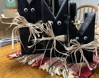May include: A set of three black wooden bunny figures with googly eyes and raffia bows. The bunnies are arranged in a row on a red and cream patterned table runner. The wooden bunnies are a decorative item.