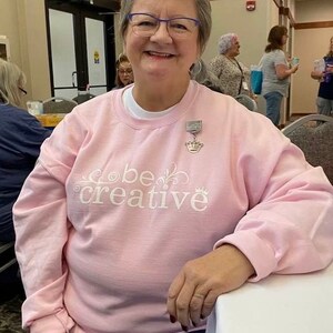 May include: A woman wearing a pink sweatshirt with the text "be creative" in white lettering. She is wearing a silver necklace with a crown charm.