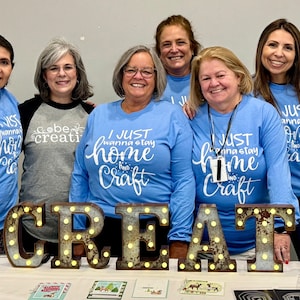 May include: Six women wearing blue long-sleeved shirts with white text that reads "I just wanna stay home and craft". They are standing in front of a sign that reads "CREATE" with light bulbs.