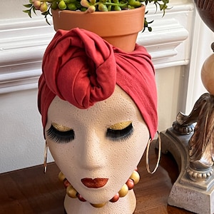 May include: A terracotta planter with a succulent plant is placed on top of a ceramic bust of a woman with a red turban. The bust has gold eyelashes, gold accents around the eyes, and a red lip. The bust is wearing a gold necklace with brown and red beads.