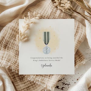 May include: A square greeting card featuring a watercolour illustration of a medal. The medal has a blue and silver ribbon and a circular emblem. Text reads, "Congratulations on being awarded the King's Ambulance Service Medal" and "Yolanda".