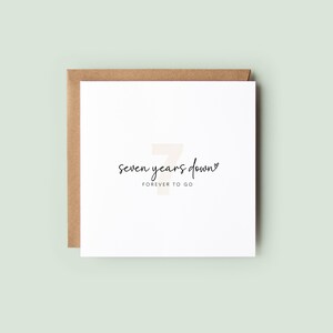 Neutral flatlay photo of a square white anniversary card with dried florals and textured fabric background. Card reads “seven years down” in script with “forever to go” below and a subtle large number 7 behind the text.