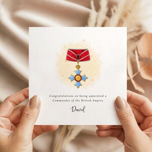 May include: A square white card featuring a watercolour illustration of a British Empire medal. The medal is red, gold, and blue. The card includes the text "Congratulations on being appointed a Commander of the British Empire" and the name "David".
