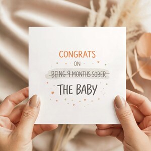 May include: A square greeting card with the text CONGRATS ON BEING 9 MONTHS SOBER THE BABY printed in orange and dark gray. The words BEING 9 MONTHS SOBER are crossed out with a line. Small dots and heart icons decorate the white card surface.