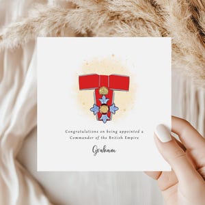 May include: A white greeting card with an illustration of a British Empire medal in red and gold. The card reads "Congratulations on being appointed a Commander of the British Empire" and is signed "Graham".
