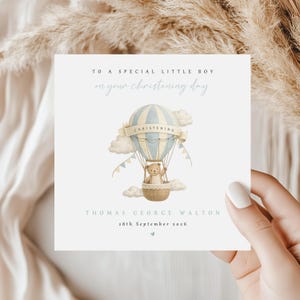 Peut inclure: Carte de baptême bleu clair et beige avec une montgolfière et un ours en peluche. Le texte dit : « À un petit garçon spécial pour votre jour de baptême » et comprend un nom et une date.