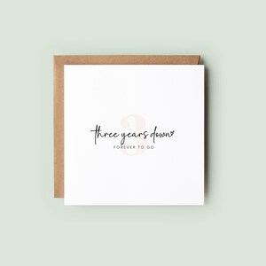 Minimalist third anniversary card showing soft neutral number 3 with text “Three Years Down” and “FOREVER TO GO”, held in hands over a neutral background with ribbon or soft fabric.