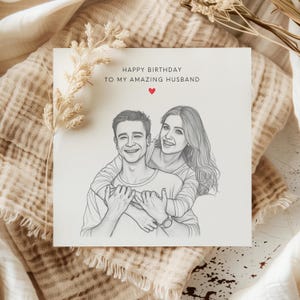 Peut inclure: Carte de vœux avec un croquis au crayon d'un couple s'embrassant. La carte porte l'inscription "HAPPY BIRTHDAY TO MY AMAZING HUSBAND" avec un cœur rouge. La carte est sur un fond de tissu beige avec des accents floraux séchés.