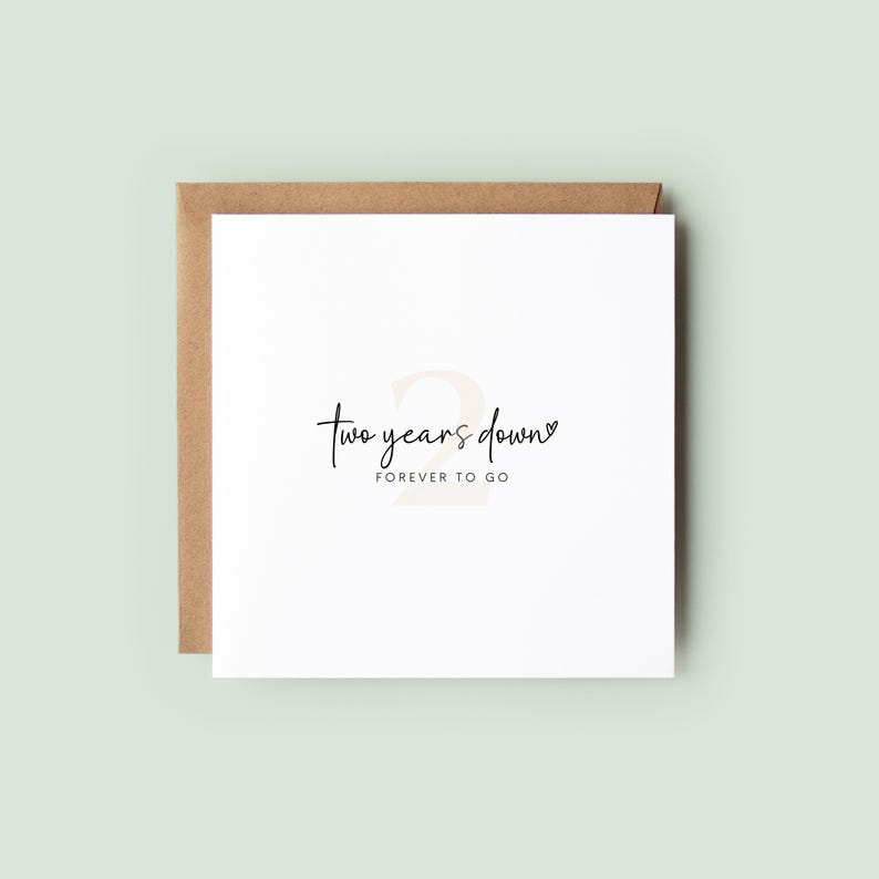 Minimalist second wedding anniversary card with a soft beige number “2” and the text “Two years down” and “FOREVER TO GO”, photographed on a white square card on a heart patterned background with ribbon in neutral tones.