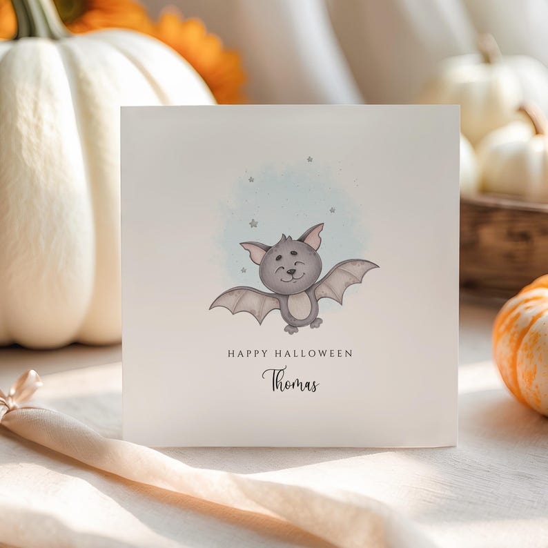 Personalised Halloween card featuring a cute smiling watercolour bat with a custom name underneath and the words "Happy Halloween" on a white greeting card.
