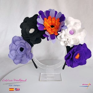 Op de afbeelding: Een haarband van vilt met bloemen in paars, wit, zwart en oranje. De haarband staat op een transparante standaard. De tekst "Catrina Headband PDF Pattern" staat onder de haarband.
