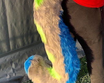 May include: A long, plush faux fur tail featuring a color-blocked pattern of vibrant lime green, soft grey, and bright blue. The tail is attached to the waist of a red long-sleeved shirt and black pants, curving downward in a distinct S-shape.