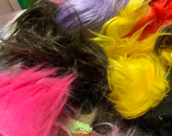 May include: A collection of faux fur scraps in various vibrant colors including bright pink, dark brown, purple, yellow, and red. The synthetic fibers are long, shaggy, and textured, appearing as a jumbled assortment of loose material pieces.