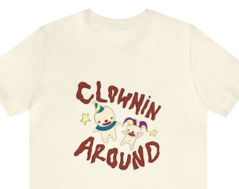 May include: A cream-coloured t-shirt with the words "Clownin Around" in a brown, whimsical font. The design features two cartoon clown figures with star accents. One clown wears a blue scarf and a pointed hat, the other a jester's hat.