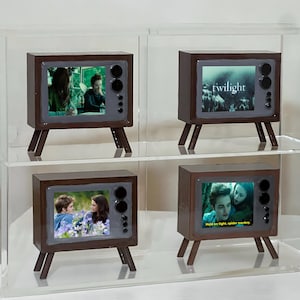 Puede incluir: Cuatro televisores marrones en miniatura con pantallas que muestran escenas de la película "Twilight". Cada televisor tiene un marco gris oscuro, detalles de altavoces negros y pequeñas patas de madera. Una pantalla muestra la palabra "twilight".