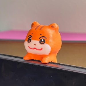 Puede incluir: Pequeña figura naranja impresa en 3D de un hámster de dibujos animados. El hámster tiene una cara blanca, ojos negros y una boca roja. La figura está posada sobre una superficie negra.