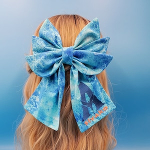 May include: A large blue hair bow with a watercolor-style pattern. The bow features a design with the word "Sivako" embroidered in orange. The bow is attached to blonde hair against a blue background.