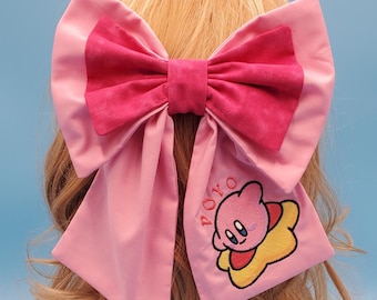 May include: A large pink bow with a smaller, darker pink bow layered on top. The bow features a cartoon character embroidery with the word "POYO". The bow is likely a hair accessory.