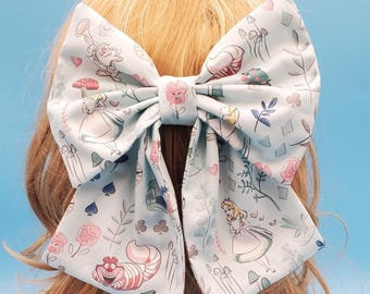 Op de afbeelding: Een grote, lichtblauwe haarstrik met een Alice in Wonderland-print. De strik toont illustraties van Alice, de Cheshire Cat, paddenstoelen en bloemelementen in roze, wit en groen. De strik is vastgebonden en op blond haar geplaatst.