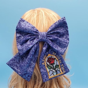 Könnte beinhalten: Eine große, blaue Stoff-Haarschleife mit einem strukturierten Muster. Die Schleife hat einen gestickten Aufnäher mit einer roten Rose in einem Glaskasten und dem Text "A Tale as old as Time".