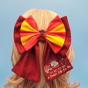 May include: A large hair bow featuring alternating red and yellow fabric panels with a central black skull and crossbones emblem. The long fabric tails are dark red with embroidered pirate ship and hat icons and the text YO HO YO HO A PIRATE'S LIFE FOR ME.