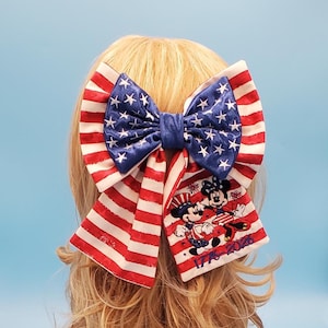 Op de afbeelding: Een grote haarstrik met een patriottisch ontwerp. De strik heeft rood-witte strepen en een blauw gedeelte met witte sterren. Het heeft een geborduurd beeld van Mickey en Minnie Mouse met de tekst "1776-2026".