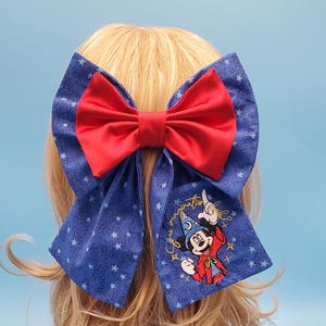 以下が含まれることがあります： 赤いリボンと白い星で飾られた青いテールが特徴の大きなヘアボウ。片方のテールには、魔法使いの衣装を着たミッキーマウスの刺繍画像と「Imagination」の文字が施されています。