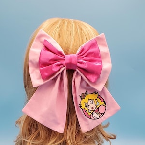 Puede incluir: Un gran lazo rosa para el cabello con un lazo rosa más oscuro encima. El lazo tiene largas colas fluidas, una de las cuales presenta una imagen bordada de la Princesa Peach de la serie de videojuegos Super Mario.