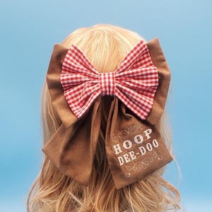 May include: A large hair bow featuring a red and white gingham bow atop a brown fabric base. The bow has long, flowing tails with the embroidered text "HOOP DEE-DOO" in white. The bow is set against a light blue background.
