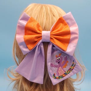 May include: A large hair bow featuring a layered design with shimmering light purple and bright orange fabric. The right tail of the bow displays an embroidered purple dragon character positioned in front of a globe, accented by a colourful rainbow arc.