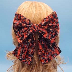 May include: A large, fabric hair bow with a floral pattern in shades of red, orange, and black. The bow is tied in the center and attached to a hair clip. The background is a light blue.