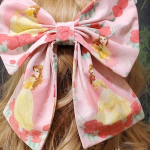 Könnte beinhalten: Eine große rosa Haarschleife mit einem sich wiederholenden Muster einer Prinzessin in einem gelben Kleid und roten Rosen. Die Schleife ist in der Mitte gebunden und vor einem Hintergrund aus blondem Haar platziert.
