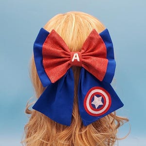 Puede incluir: Un lazo grande para el cabello con un centro rojo brillante y la letra blanca "A". El lazo tiene detalles en azul y un emblema circular con una estrella blanca. Este accesorio es ideal para eventos temáticos.