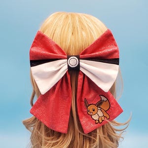 Peut inclure: Un grand nœud rouge, blanc et noir avec un personnage Eevee brodé. Le nœud est attaché à une perruque blonde. Le nœud est en tissu rouge texturé avec un centre blanc et une bande noire. Le personnage Eevee est marron, beige et blanc.