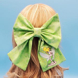 May include: A large, lime green hair bow with a glittery, watercolour-like pattern. The bow features an embroidered image of Tinkerbell on one side. The bow is attached to a blonde wig, set against a light blue background.