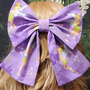 Könnte beinhalten: Eine große lila Haarschleife mit einem sich wiederholenden Muster aus Tinkerbell-Illustrationen und Schmetterlingen. Die Schleife ist aus Stoff und in Form einer großen Schleife gebunden. Das Wort "Bell" ist auf der Schleife aufgedruckt.