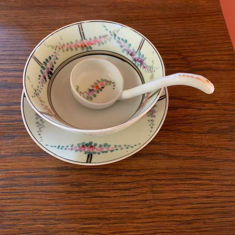 May include: A white porcelain soup bowl with a floral design and a matching ladle. The bowl and ladle are decorated with a delicate floral pattern in shades of pink, purple, and green.