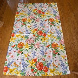 May include: A white tablecloth with a floral pattern featuring yellow, orange, pink, and blue flowers. The tablecloth has a white background and a green border.