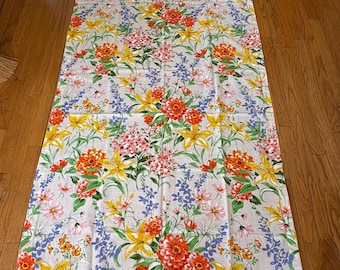 May include: A white tablecloth with a floral pattern featuring yellow, orange, pink, and blue flowers. The tablecloth has a white background and a green border.