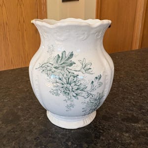 Op de afbeelding: Witte keramische vaas met een groen bloemenontwerp. De vaas heeft een gegolfde rand en een rond lichaam met een reliëfdesign.